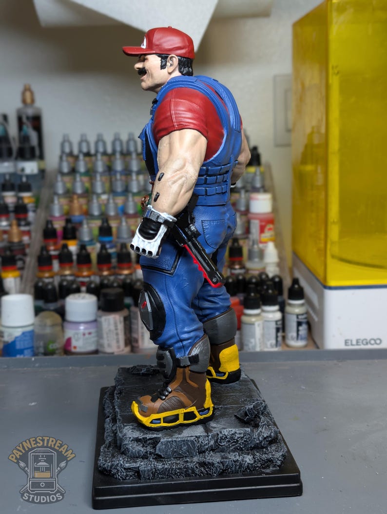 May include: A detailed figurine of a character in a red cap, red shirt, and blue overalls, with a mustache. The figure is standing on a gray base, with a black gun and white gloves. The figure is a stylized representation of a video game character.