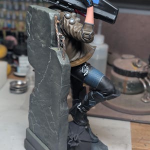 May include: A detailed figurine of a character leaning against a gray concrete pillar, holding a large black rifle with a red top section. The character wears a brown jacket, black pants with blue ribbed sections, and black boots, with a small logo at the base.