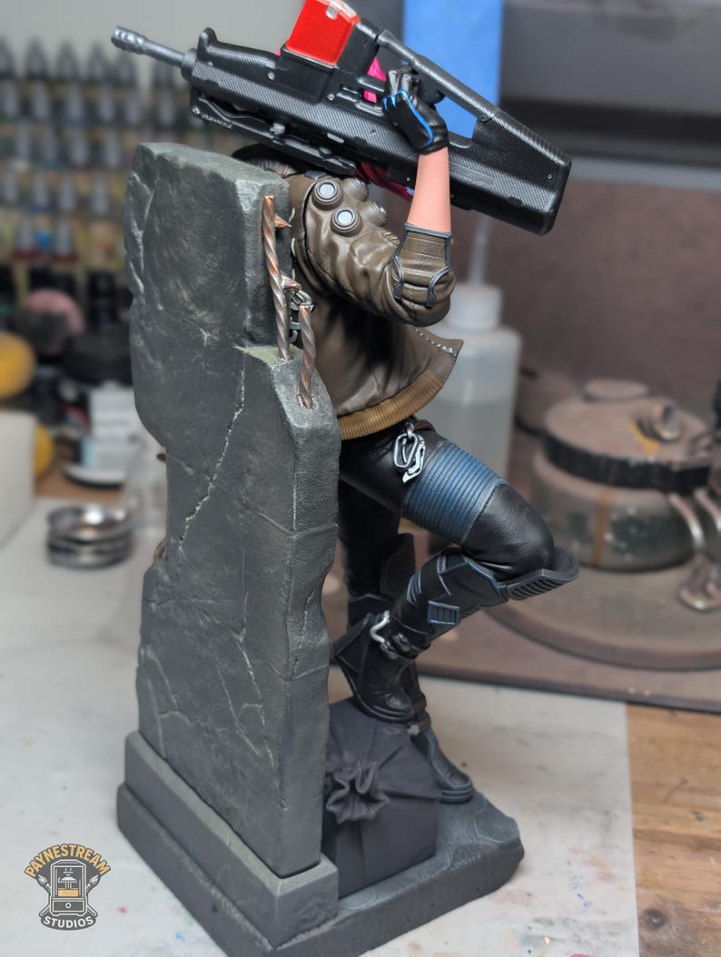 May include: A detailed figurine of a character leaning against a gray concrete pillar, holding a large black rifle with a red top section. The character wears a brown jacket, black pants with blue ribbed sections, and black boots, with a small logo at the base.