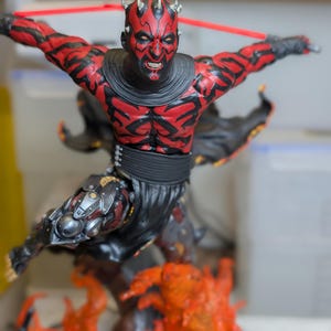 K&ouml;nnte beinhalten: Eine detaillierte Figur eines rot-schwarzen geh&ouml;rnten Charakters mit Gesichtst&auml;towierungen, bekleidet mit einer schwarzen Tunika und einem G&uuml;rtel. Die Figur steht in dynamischer Pose &uuml;ber einem orange-gelben Flammensockel und h&auml;lt eine rote Waffe.