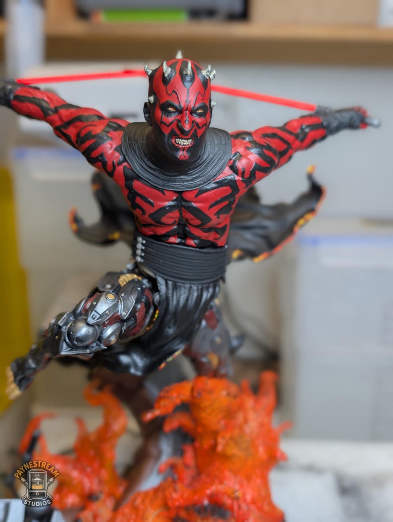 K&ouml;nnte beinhalten: Eine detaillierte Figur eines rot-schwarzen geh&ouml;rnten Charakters mit Gesichtst&auml;towierungen, bekleidet mit einer schwarzen Tunika und einem G&uuml;rtel. Die Figur steht in dynamischer Pose &uuml;ber einem orange-gelben Flammensockel und h&auml;lt eine rote Waffe.
