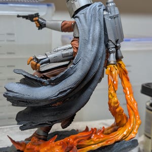 K&ouml;nnte beinhalten: Detaillierte Figur einer Figur aus einer Science-Fiction-Serie. Die Figur tr&auml;gt einen silbernen Helm, eine braune und graue R&uuml;stung und einen grauen Umhang. Die Figur schie&szlig;t mit einer schwarzen Pistole, wobei orangefarbene Flammen aus dem Jetpack und der Basis austreten.