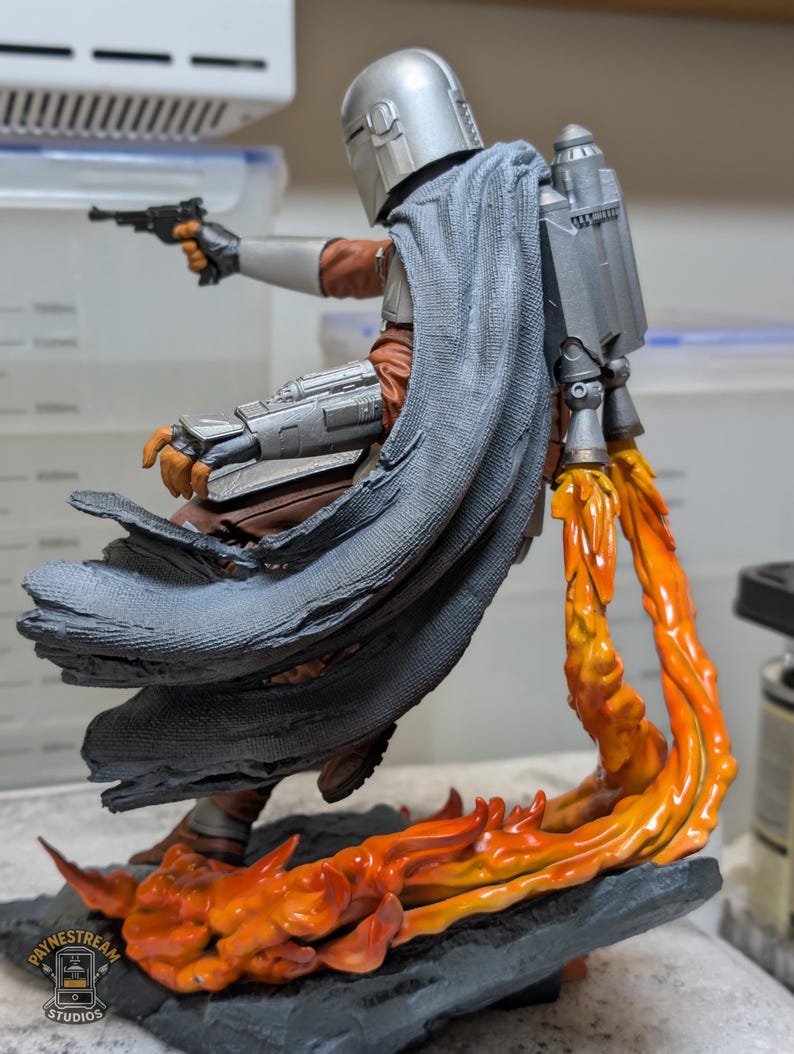 K&ouml;nnte beinhalten: Detaillierte Figur einer Figur aus einer Science-Fiction-Serie. Die Figur tr&auml;gt einen silbernen Helm, eine braune und graue R&uuml;stung und einen grauen Umhang. Die Figur schie&szlig;t mit einer schwarzen Pistole, wobei orangefarbene Flammen aus dem Jetpack und der Basis austreten.
