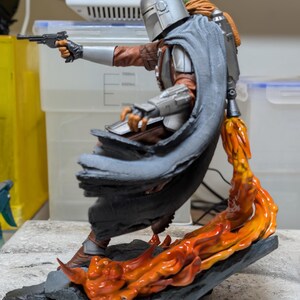 K&ouml;nnte beinhalten: Detaillierte Figur von The Mandalorian in dynamischer Pose, die eine Blasterpistole h&auml;lt. Die Figur ist mit silberner R&uuml;stung, einer braunen Jacke und einem grauen Umhang bekleidet. Orangefarbene Flammen schlagen aus der Basis und erzeugen einen dramatischen Effekt.
