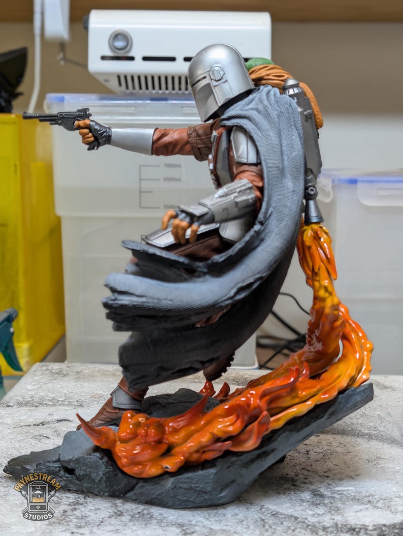 K&ouml;nnte beinhalten: Detaillierte Figur von The Mandalorian in dynamischer Pose, die eine Blasterpistole h&auml;lt. Die Figur ist mit silberner R&uuml;stung, einer braunen Jacke und einem grauen Umhang bekleidet. Orangefarbene Flammen schlagen aus der Basis und erzeugen einen dramatischen Effekt.