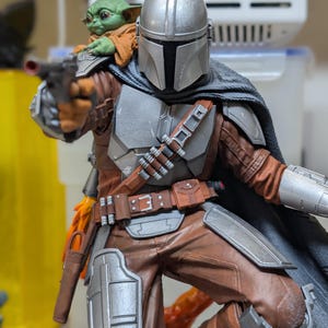 K&ouml;nnte beinhalten: Detaillierte Figur von The Mandalorian, in silberner R&uuml;stung und brauner Kleidung, mit einem Blaster. Eine kleine, gr&uuml;n geh&auml;utete Grogu-Figur sitzt auf seiner Schulter. Die Figur ist dynamisch posiert, mit einem schwarzen Umhang und einem detaillierten G&uuml;rtel.