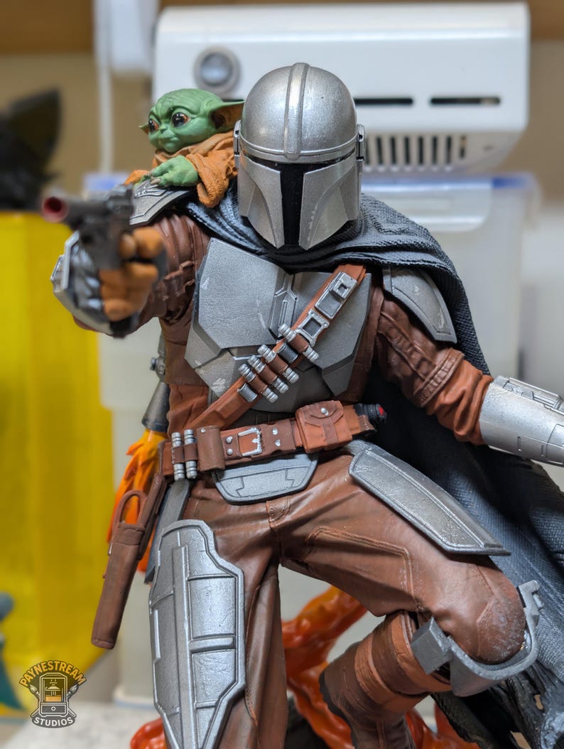 K&ouml;nnte beinhalten: Detaillierte Figur von The Mandalorian, in silberner R&uuml;stung und brauner Kleidung, mit einem Blaster. Eine kleine, gr&uuml;n geh&auml;utete Grogu-Figur sitzt auf seiner Schulter. Die Figur ist dynamisch posiert, mit einem schwarzen Umhang und einem detaillierten G&uuml;rtel.