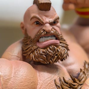 Puede incluir: Una figura detallada de un personaje musculoso con peinado mohawk, una barba marr&oacute;n espesa y una expresi&oacute;n facial agresiva que muestra los dientes. La figura tiene piel bronceada, hombros anchos y una prenda texturizada de color marr&oacute;n.