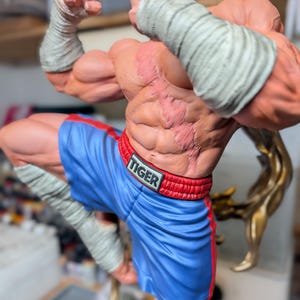May include: A muscular figurine of a fighter with a prominent scar across the chest, wearing blue boxing shorts with a red waistband labeled TIGER. The figure has white hand wraps on the wrists and lower legs, posed in a dynamic stance.