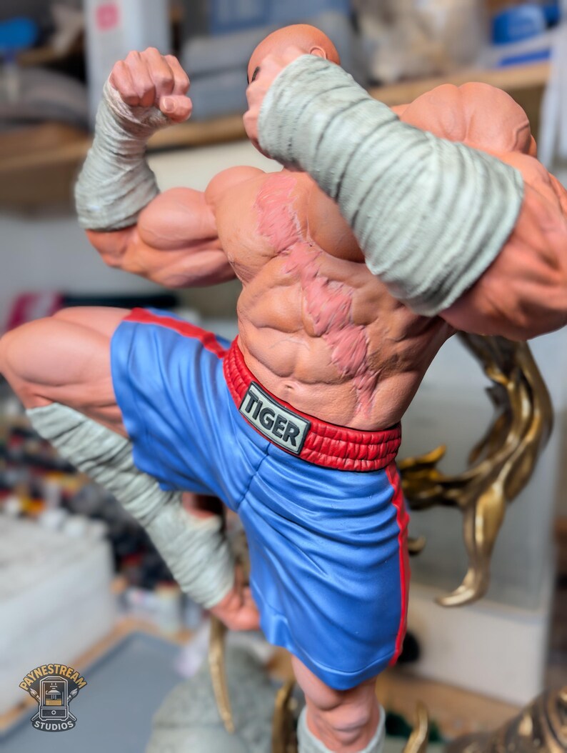 May include: A muscular figurine of a fighter with a prominent scar across the chest, wearing blue boxing shorts with a red waistband labeled TIGER. The figure has white hand wraps on the wrists and lower legs, posed in a dynamic stance.
