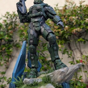 May include: A Master Chief action figure in olive green armor with a gold visor, holding a rifle. The figure stands on a rocky base with blue geometric structures and green grass. A small logo for Paynestream Studios is visible in the corner.