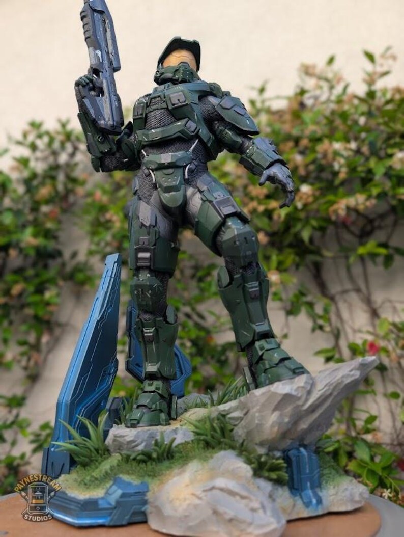 May include: A Master Chief action figure in olive green armor with a gold visor, holding a rifle. The figure stands on a rocky base with blue geometric structures and green grass. A small logo for Paynestream Studios is visible in the corner.