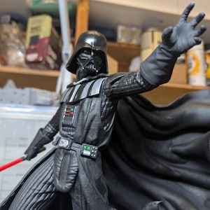 May include: A Darth Vader action figure in a black suit with a helmet, cape, and chest control panel featuring red and green buttons. The figure has one arm extended forward with an open hand and holds a red lightsaber in the other hand.