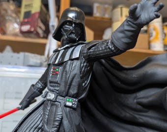 May include: A Darth Vader action figure in a black suit with a helmet, cape, and chest control panel featuring red and green buttons. The figure has one arm extended forward with an open hand and holds a red lightsaber in the other hand.