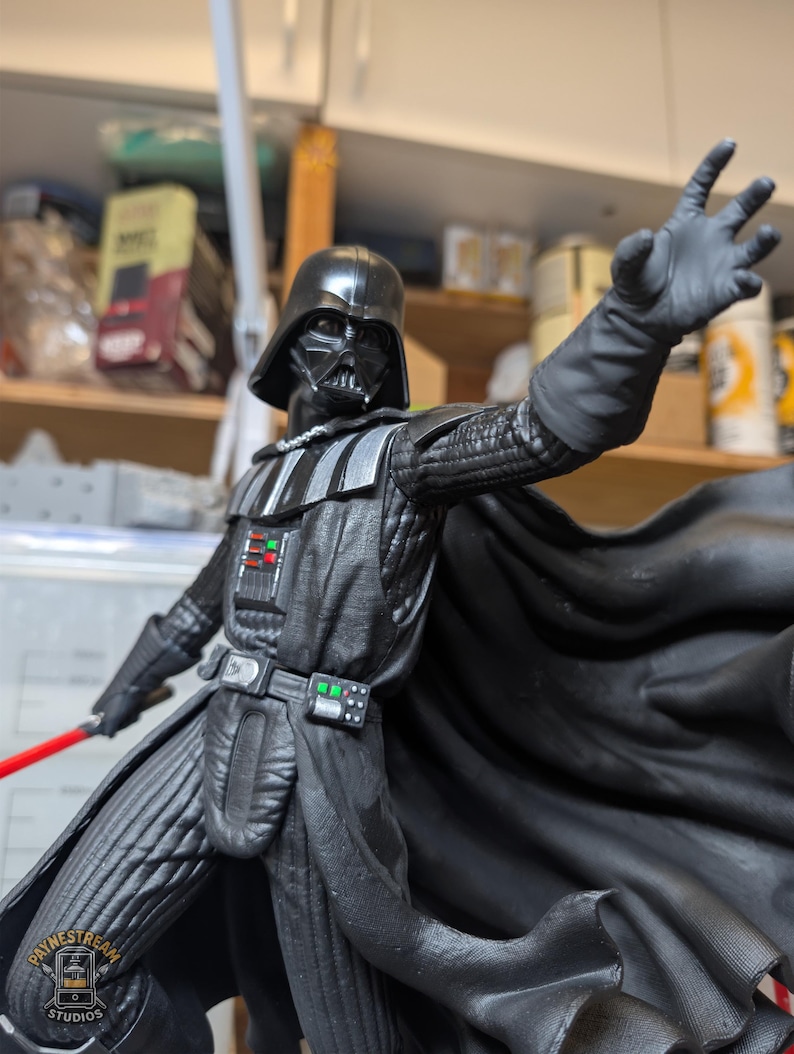 May include: A Darth Vader action figure in a black suit with a helmet, cape, and chest control panel featuring red and green buttons. The figure has one arm extended forward with an open hand and holds a red lightsaber in the other hand.