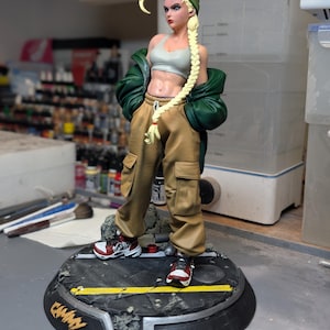 May include: A collectible figurine of a blonde character with a long braid wearing a green beanie, a light gray sports bra, a dark green jacket, and tan cargo pants. The figure stands on a circular base with a street design and the text CAMMY.