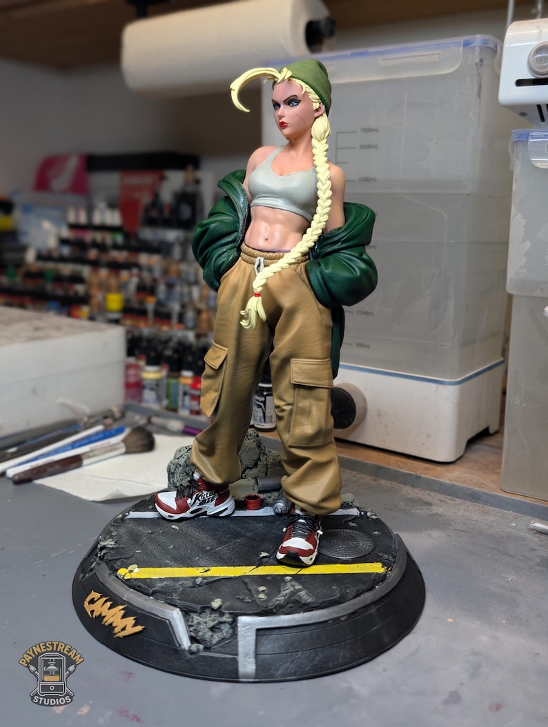 May include: A collectible figurine of a blonde character with a long braid wearing a green beanie, a light gray sports bra, a dark green jacket, and tan cargo pants. The figure stands on a circular base with a street design and the text CAMMY.