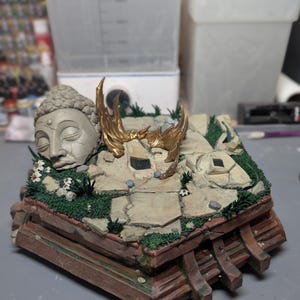 May include: A diorama base featuring a weathered gray Buddha head sculpture, metallic gold wing accents, and cracked stone flooring with patches of green moss and small white flowers. The base has a brown, tiered wooden-style frame structure.