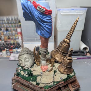 May include: A painted resin sculpture featuring a single human leg wearing blue boxing shorts with the word TIGER on the waistband. The leg is wrapped in white bandages and stands on a stone base with a stone Buddha head and two gold bells.