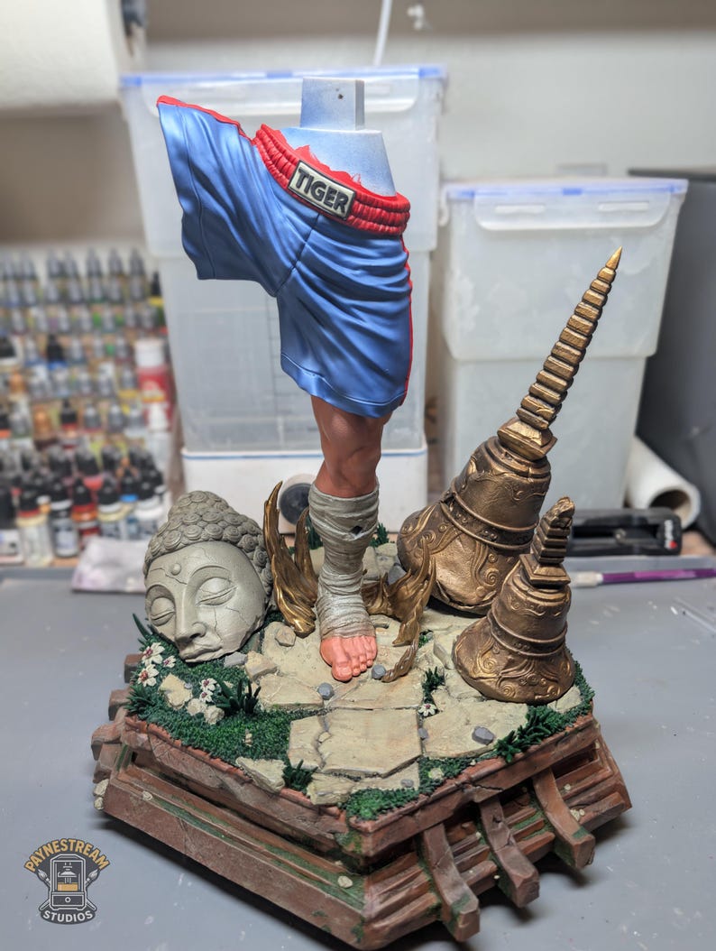 May include: A painted resin sculpture featuring a single human leg wearing blue boxing shorts with the word TIGER on the waistband. The leg is wrapped in white bandages and stands on a stone base with a stone Buddha head and two gold bells.