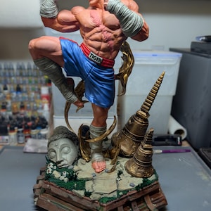 May include: A muscular, bald figurine in a fighting pose, wearing blue shorts with the word TIGER on the waistband and white hand wraps. The figure stands on a base featuring a stone Buddha head, golden bells, and green foliage, with a logo for Paynestream Studios in the corner.