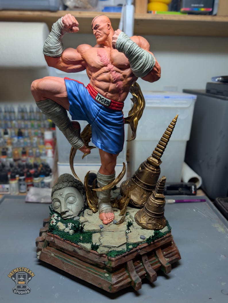 May include: A muscular, bald figurine in a fighting pose, wearing blue shorts with the word TIGER on the waistband and white hand wraps. The figure stands on a base featuring a stone Buddha head, golden bells, and green foliage, with a logo for Paynestream Studios in the corner.