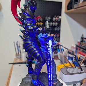 May include: A detailed figurine featuring a dark armored warrior with a vibrant red ponytail and glowing blue accents, standing on a rocky base. The warrior holds a large, luminous blue sword with intricate white patterns, positioned behind a person.