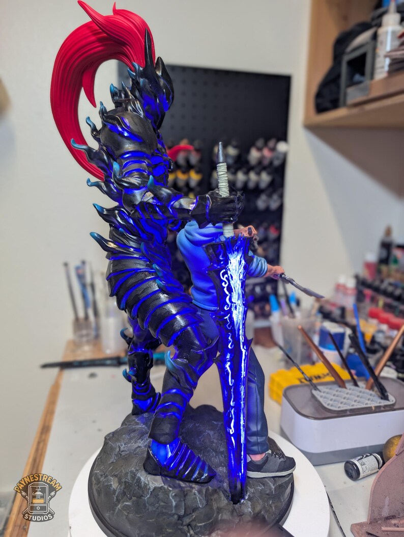 May include: A detailed figurine featuring a dark armored warrior with a vibrant red ponytail and glowing blue accents, standing on a rocky base. The warrior holds a large, luminous blue sword with intricate white patterns, positioned behind a person.