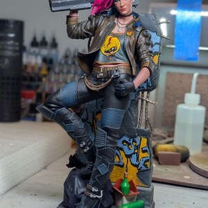 May include: A detailed collectible figurine of a character with bright pink hair, wearing a brown jacket over a gray graphic t-shirt, black pants, and boots. The figure holds a large futuristic rifle and stands against a graffiti-covered wall base.