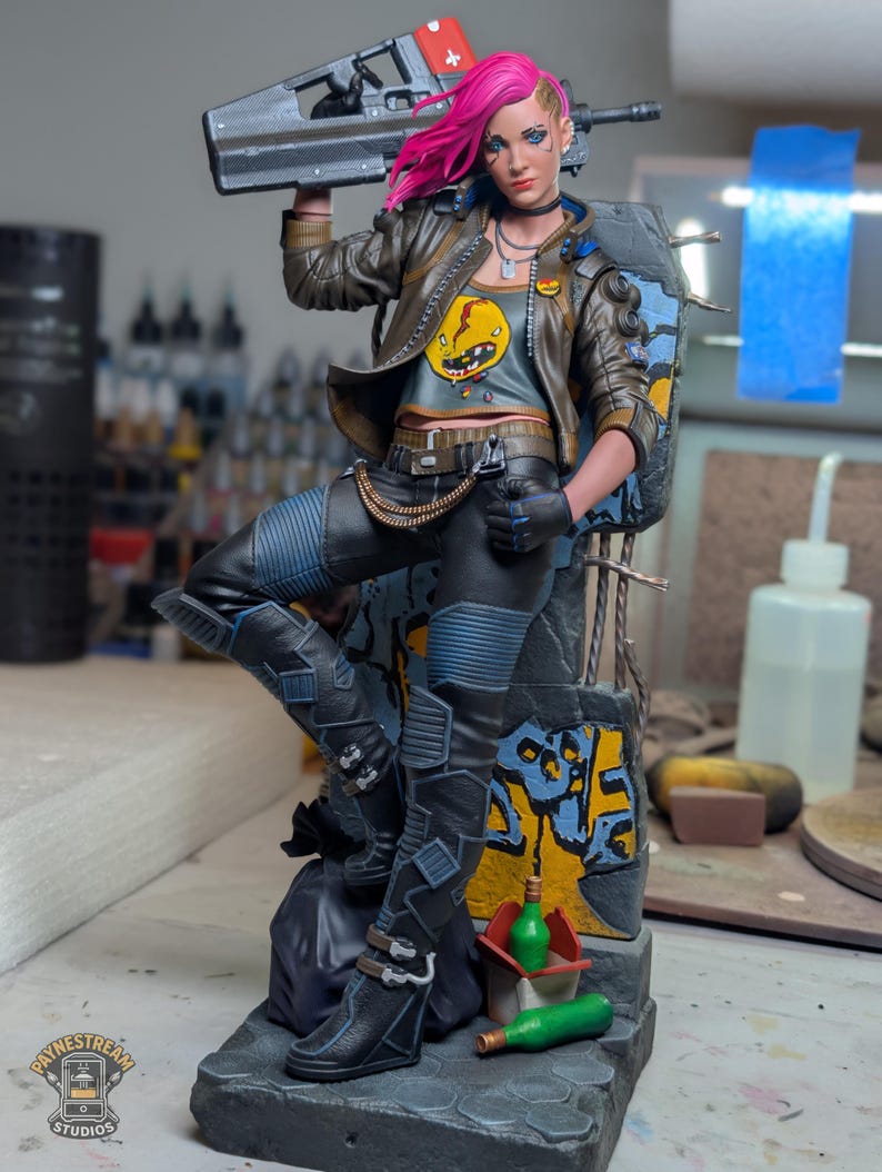 May include: A detailed collectible figurine of a character with bright pink hair, wearing a brown jacket over a gray graphic t-shirt, black pants, and boots. The figure holds a large futuristic rifle and stands against a graffiti-covered wall base.
