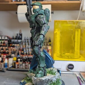 May include: A detailed green Master Chief figurine from Halo, standing on a rocky base with green grass. The character wears iconic green armor with a gold visor and holds a rifle. A logo for Paynestream Studios is visible in the bottom left corner.