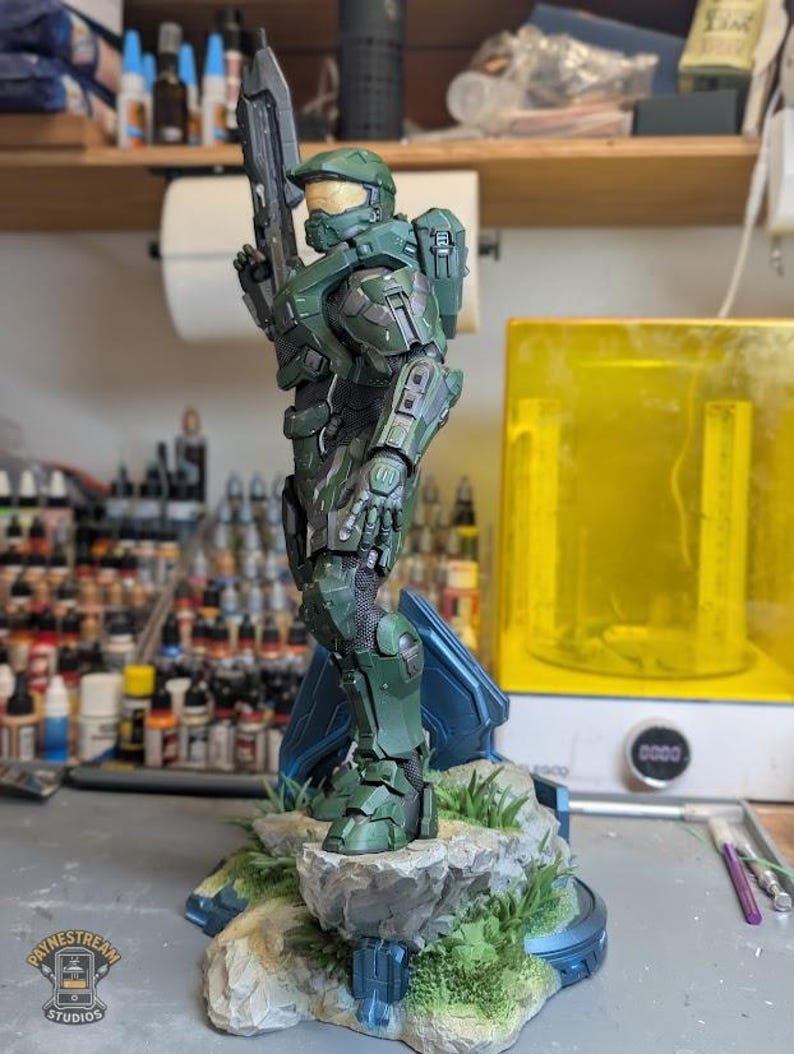May include: A detailed green Master Chief figurine from Halo, standing on a rocky base with green grass. The character wears iconic green armor with a gold visor and holds a rifle. A logo for Paynestream Studios is visible in the bottom left corner.