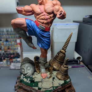 May include: A muscular, bald male figurine in a dynamic fighting pose, wearing blue shorts with a red waistband labeled TIGER. The figure has a prominent chest scar, armless shoulders, and leg wraps. It stands on a base with a stone head and bells.