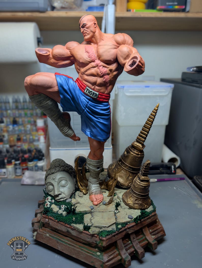 May include: A muscular, bald male figurine in a dynamic fighting pose, wearing blue shorts with a red waistband labeled TIGER. The figure has a prominent chest scar, armless shoulders, and leg wraps. It stands on a base with a stone head and bells.