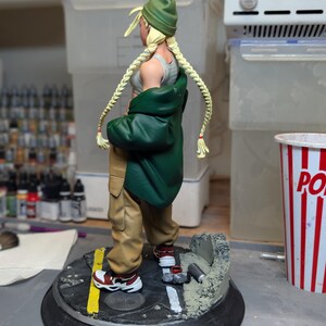 May include: A collectible figurine of a character with blonde braided hair wearing a green beanie, a grey tank top, a dark green jacket, and tan cargo pants. The figure stands on a circular base designed like a street with a yellow line and debris.