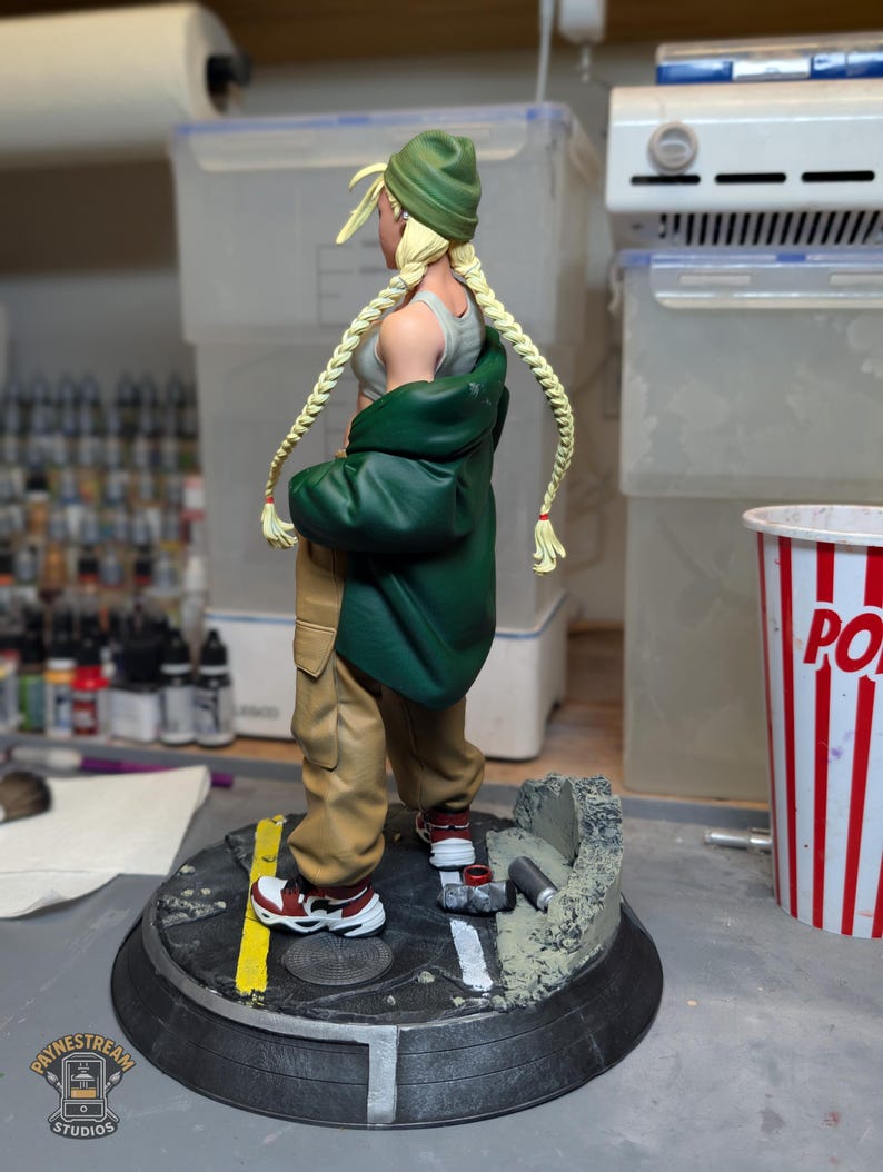 May include: A collectible figurine of a character with blonde braided hair wearing a green beanie, a grey tank top, a dark green jacket, and tan cargo pants. The figure stands on a circular base designed like a street with a yellow line and debris.