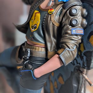 May include: A collectible figurine wearing a brown leather-style jacket with blue accents, circular shoulder details, and a patch. The figure wears a dark gray tank top with a yellow graphic, black pants, and a black glove with blue trim.
