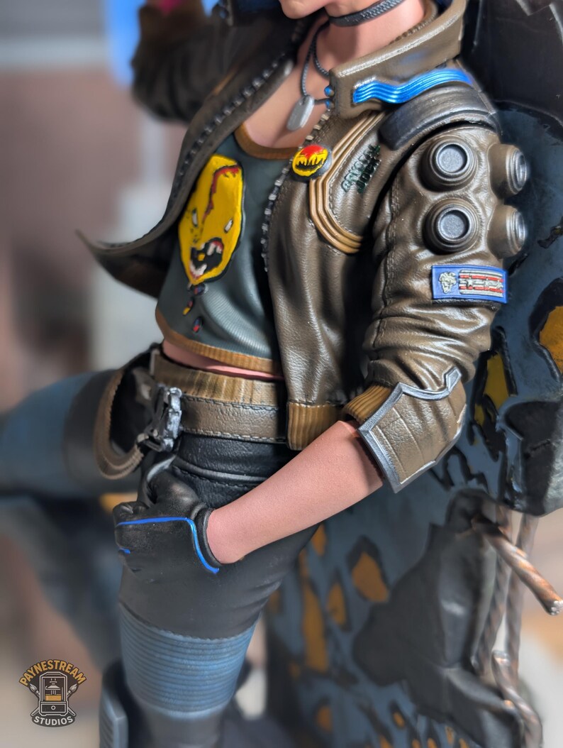 May include: A collectible figurine wearing a brown leather-style jacket with blue accents, circular shoulder details, and a patch. The figure wears a dark gray tank top with a yellow graphic, black pants, and a black glove with blue trim.