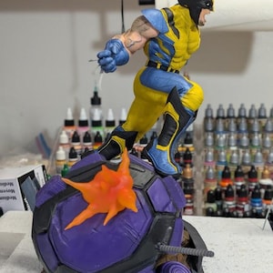 May include: A detailed figurine of Wolverine in a dynamic pose, clad in his classic yellow and blue costume. The figure is leaping from a purple and black base with an orange explosion effect. The base has the text "PAYNESTREAM STUDIOS".