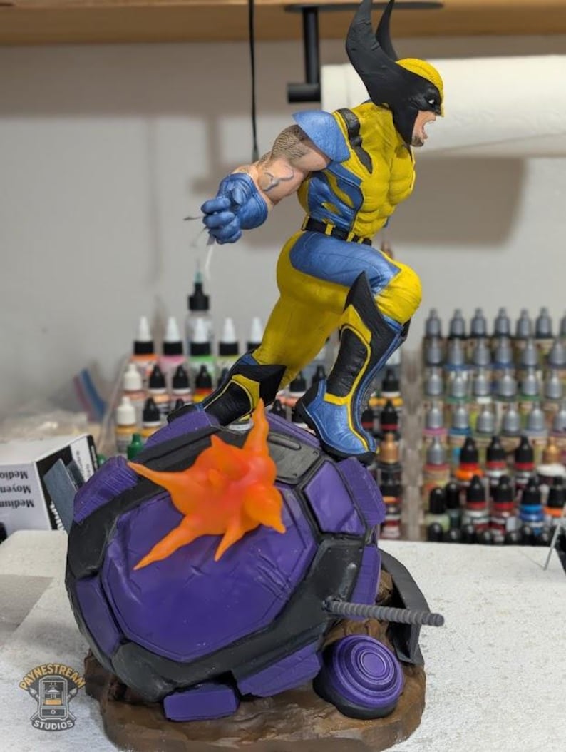 May include: A detailed figurine of Wolverine in a dynamic pose, clad in his classic yellow and blue costume. The figure is leaping from a purple and black base with an orange explosion effect. The base has the text "PAYNESTREAM STUDIOS".