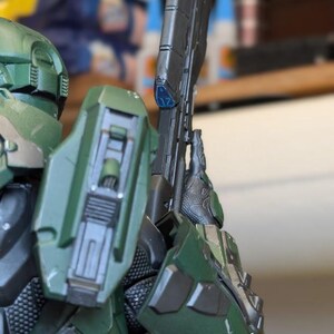 May include: A green armored action figure wearing a helmet and shoulder plating, holding a long black rifle with a blue accent piece marked with the number 17. The figure features detailed mechanical textures and a logo reading Paynestream Studios.