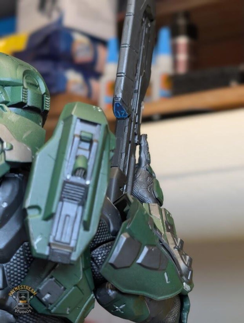 May include: A green armored action figure wearing a helmet and shoulder plating, holding a long black rifle with a blue accent piece marked with the number 17. The figure features detailed mechanical textures and a logo reading Paynestream Studios.