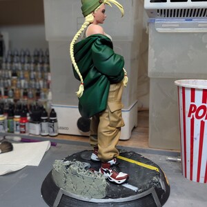 May include: A collectible figurine of a character with long blonde braided hair wearing a green beanie, a dark green oversized jacket, a white top, and tan cargo pants. The figure wears red, white, and black sneakers and stands on a circular base.