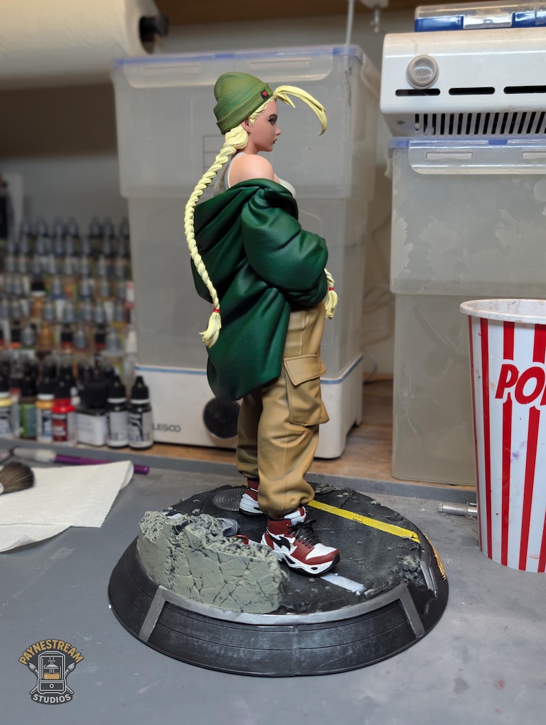 May include: A collectible figurine of a character with long blonde braided hair wearing a green beanie, a dark green oversized jacket, a white top, and tan cargo pants. The figure wears red, white, and black sneakers and stands on a circular base.