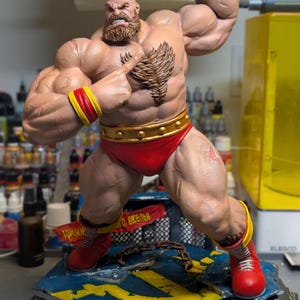 Puede incluir: Una figura musculosa de Zangief con cresta y barba, que lleva un ba&ntilde;ador rojo, mu&ntilde;equeras amarillas y botas rojas. La figura se apoya sobre una base azul y amarilla con una cadena, un estandarte rojo con texto cir&iacute;lico y el logotipo de Paynestream Studios.