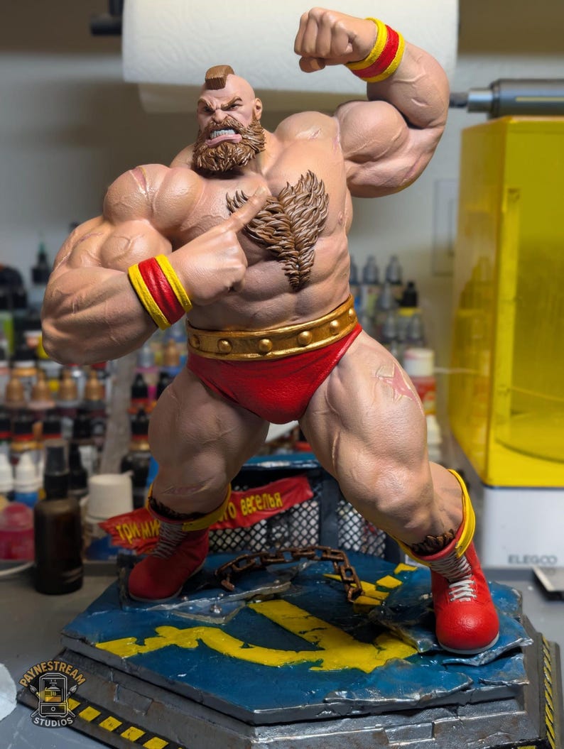 Puede incluir: Una figura musculosa de Zangief con cresta y barba, que lleva un ba&ntilde;ador rojo, mu&ntilde;equeras amarillas y botas rojas. La figura se apoya sobre una base azul y amarilla con una cadena, un estandarte rojo con texto cir&iacute;lico y el logotipo de Paynestream Studios.