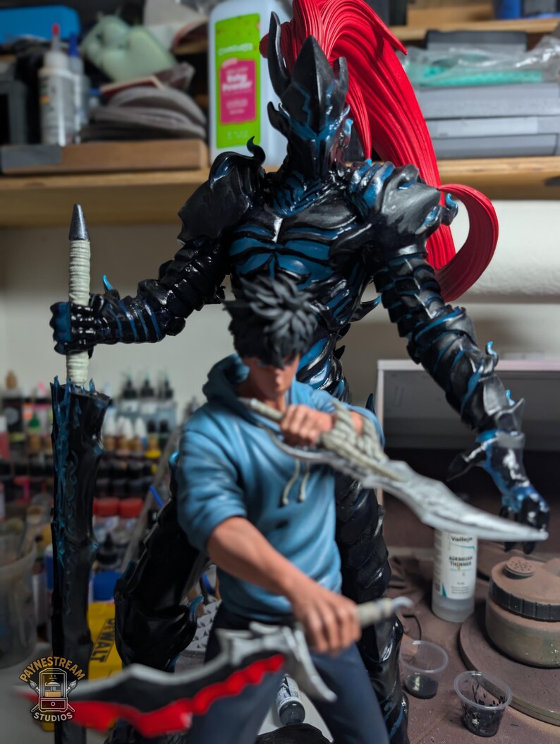 May include: A detailed figurine featuring a dark armored warrior with a long red ponytail standing behind a person in a blue hoodie. The warrior holds a tall sword, while the person in the hoodie wields two curved blades. A logo reads Paynestream Studios.