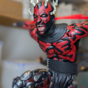 K&ouml;nnte beinhalten: Eine detaillierte Figur mit rot-schwarzen Hautmustern, gelben Augen und spitzen Z&auml;hnen. Die Figur hat helle H&ouml;rner auf dem Kopf, einen schwarzen Halskragen und metallische mechanische Beinbauteile sowie einen dunklen, strukturierten Rock.