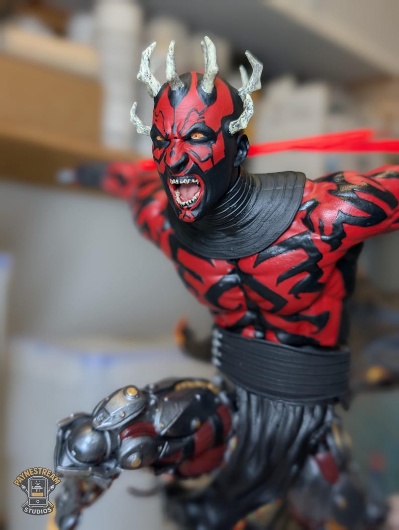 K&ouml;nnte beinhalten: Eine detaillierte Figur mit rot-schwarzen Hautmustern, gelben Augen und spitzen Z&auml;hnen. Die Figur hat helle H&ouml;rner auf dem Kopf, einen schwarzen Halskragen und metallische mechanische Beinbauteile sowie einen dunklen, strukturierten Rock.