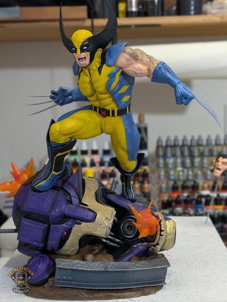 May include: A Wolverine figurine in a dynamic pose, clad in a yellow and blue costume, with claws extended. The figure stands atop a purple and gold robotic head, with fiery orange bursts. The base features the "Paynestream Studios" logo.